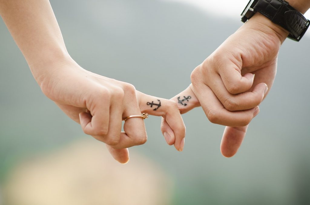 Zwei Personen mit Partner-Tattoo am Zeigefinder halten sich an der Hand. In der Nahaufnahme sind nur die Hände zu sehen.