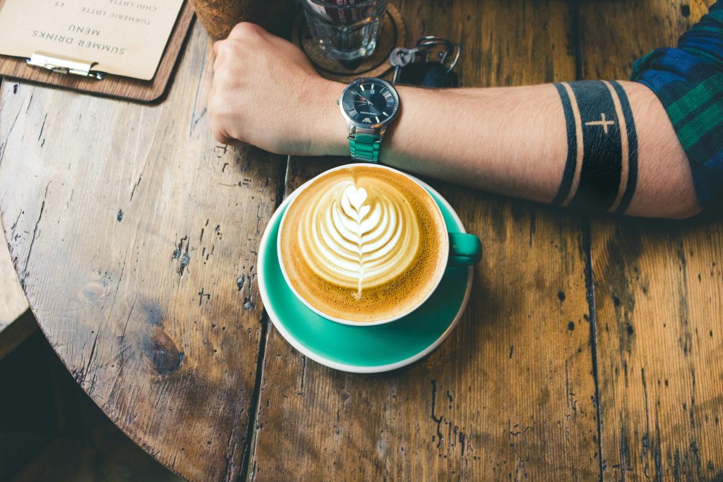 Nahaufnahme eines Unterarms mit Armband-Tattoo neben einer Tasse Cappuccino.