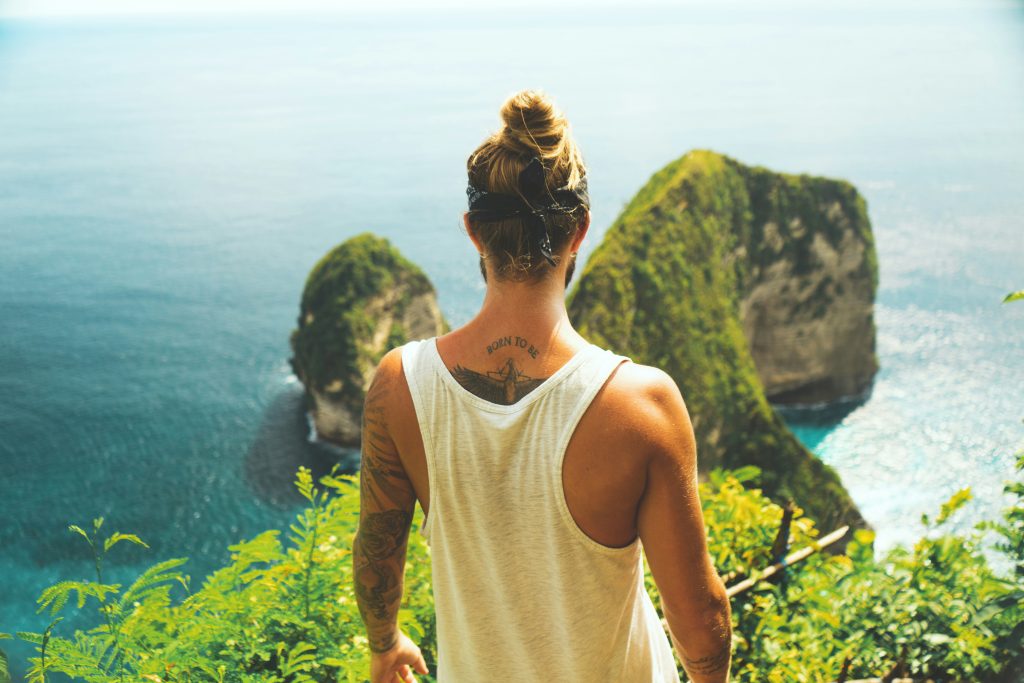 Foto eines Mannes vor Klippenkulisse und Meer. Der Mann hat auf Rücken und Armen Tattoos.