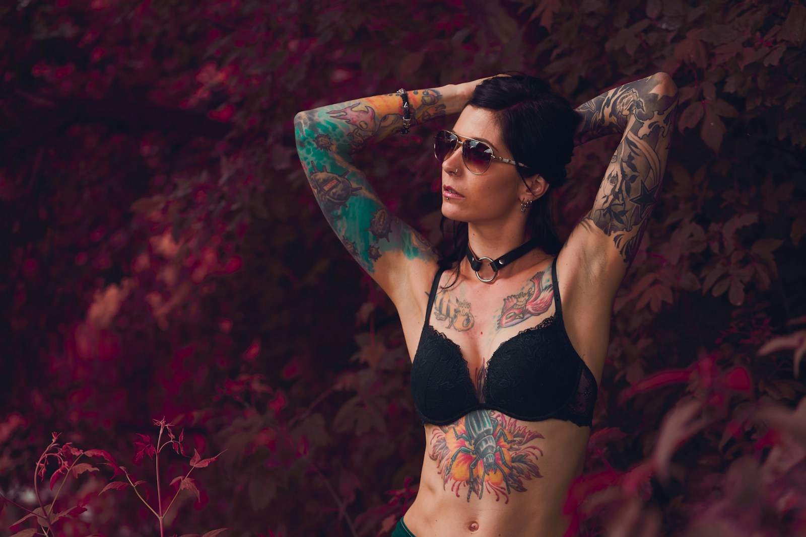 Eine Frau mit Tattoos auf den Armen steht vor einem rötlichen Hintergrund. Sie trägt einen schwarzen BH.
