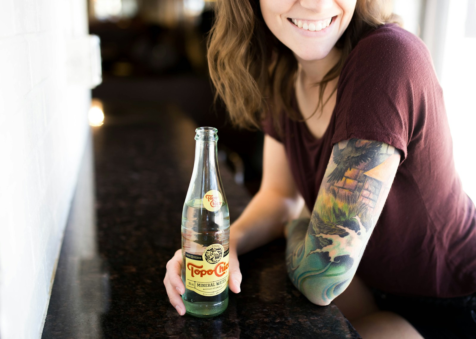 candid photography of woman holding bottle with broad smile, a big tattoo on her arm.