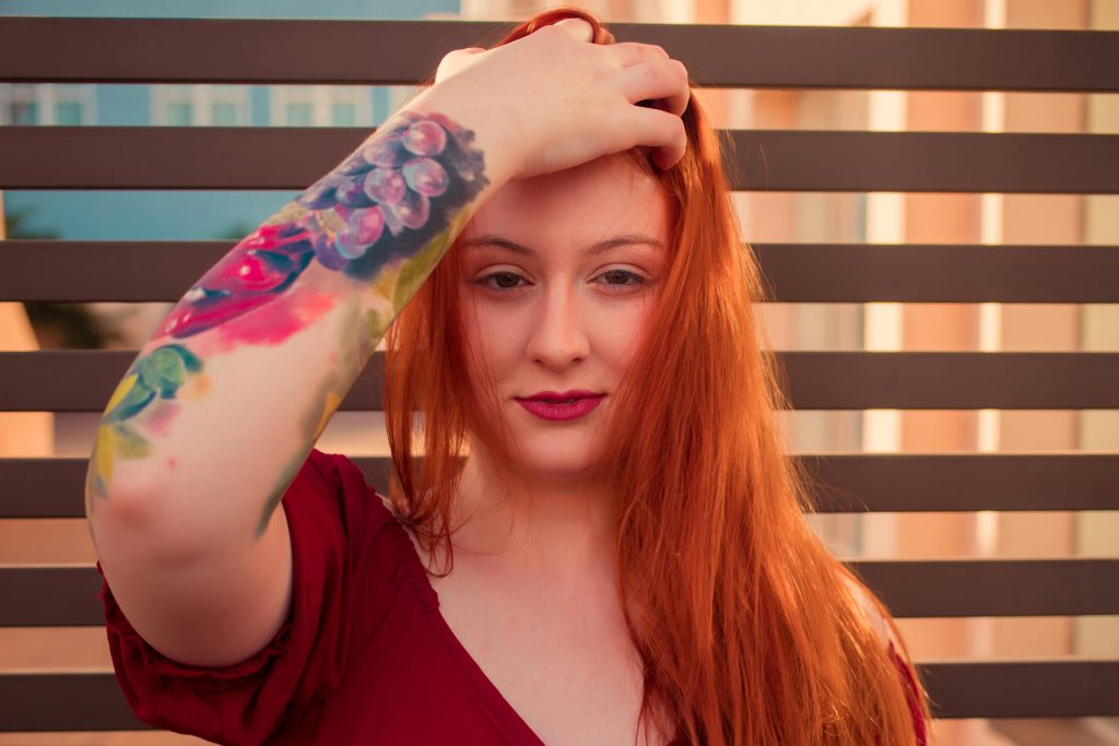Porträtfoto einer rothaarigen Frau, die sich mit der Hand durch die offenen Haare fährt. Auf ihrem Unterarm ist ein großes Watercolor-Tattoo zu sehen.