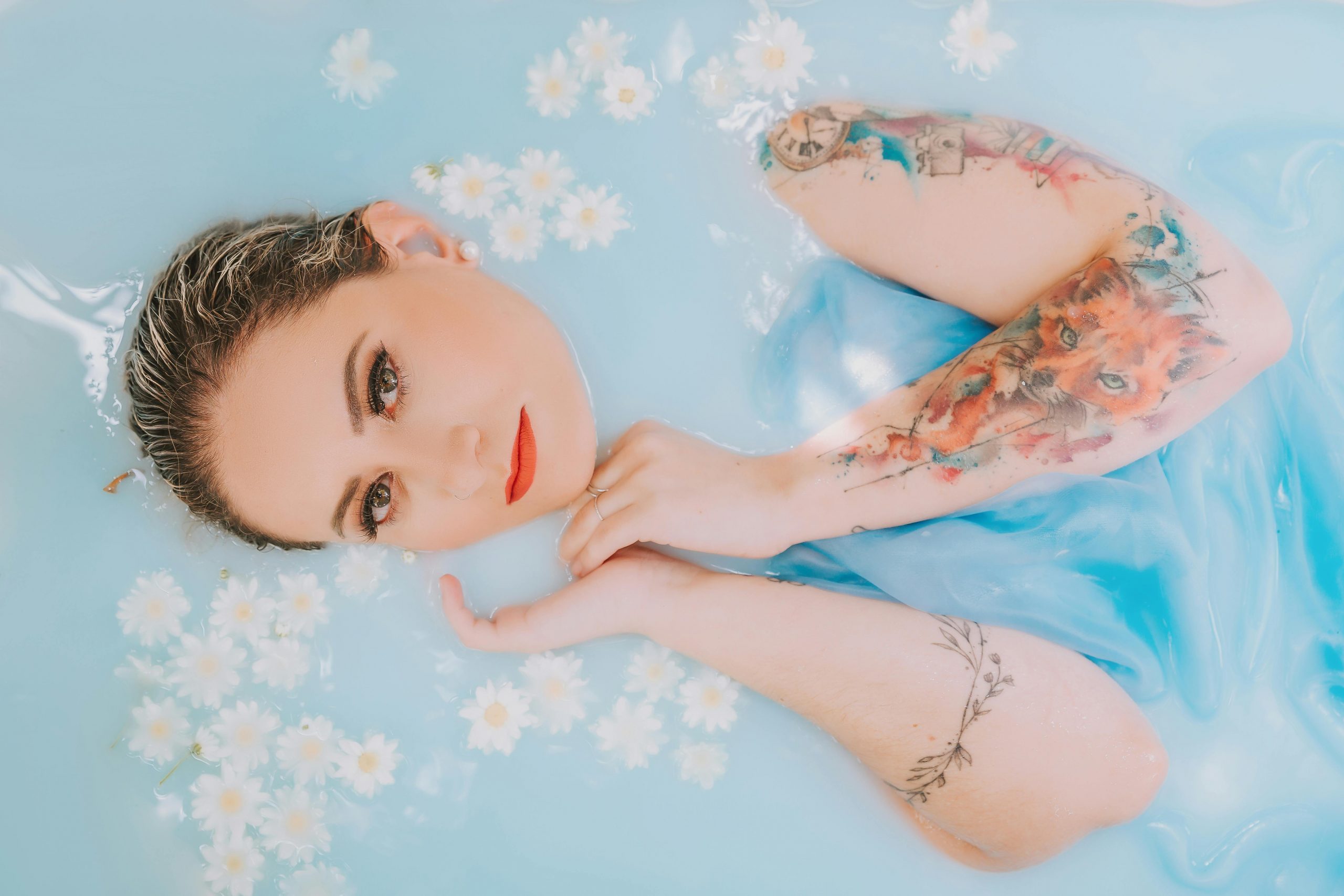 Eine Frau liegt in hellblauem Wasser, in dem weiße Blumen schwimmen. Nur ihre Arme und Gesicht sind zu sehen. Auf dem Arm sind mehrere Watercolor-Tattoos zu sehen.