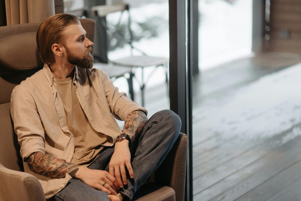 Foto eines Mannes, der vor einer Fensterfront in einem Sessel sitzt. Seine Hemdsärmel sind hochgerollt, sodass man an beiden Unterarmen Tattoos sieht.