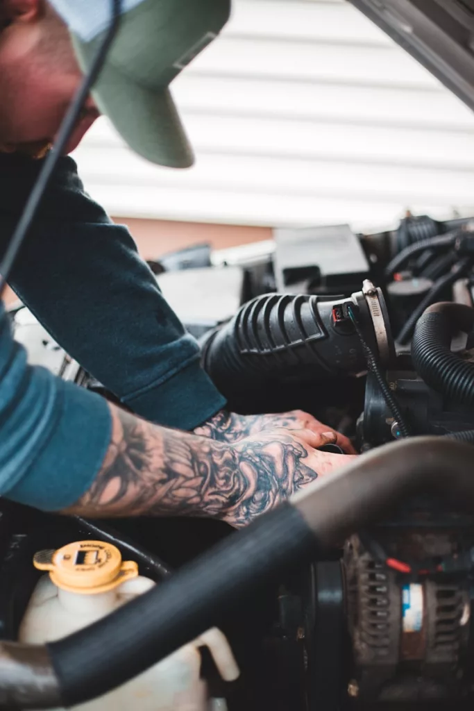Eine Person mit blauem Pullover und Tattoo auf dem Unterarm arbeitet mit den Händen in einer offenen Motorhaube.