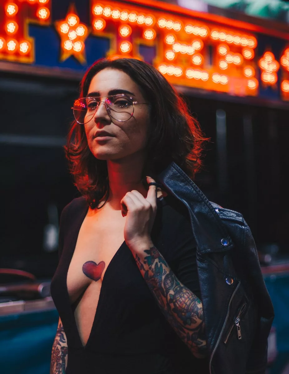 woman in black plunging-neckline holding black leather jacket. On her arms and between her breast are tattoos.