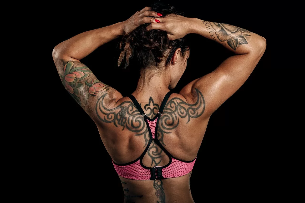 Rückenansicht einer Frau. Sie trägt einen pinken Sport-BH und hält ihre Haare mit beiden Händen nach oben. Auf Schultern, Armen und Wirbelsäule sind Tattoos zu sehen.