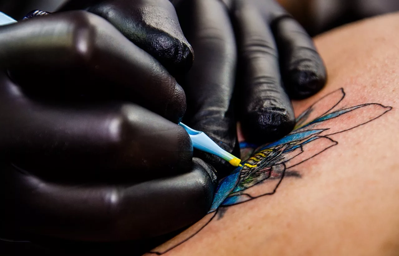Nahaufnahme von zwei Händen. Eine Person sticht ein Blumen-Tattoo, das bunt eingefärbt wird.