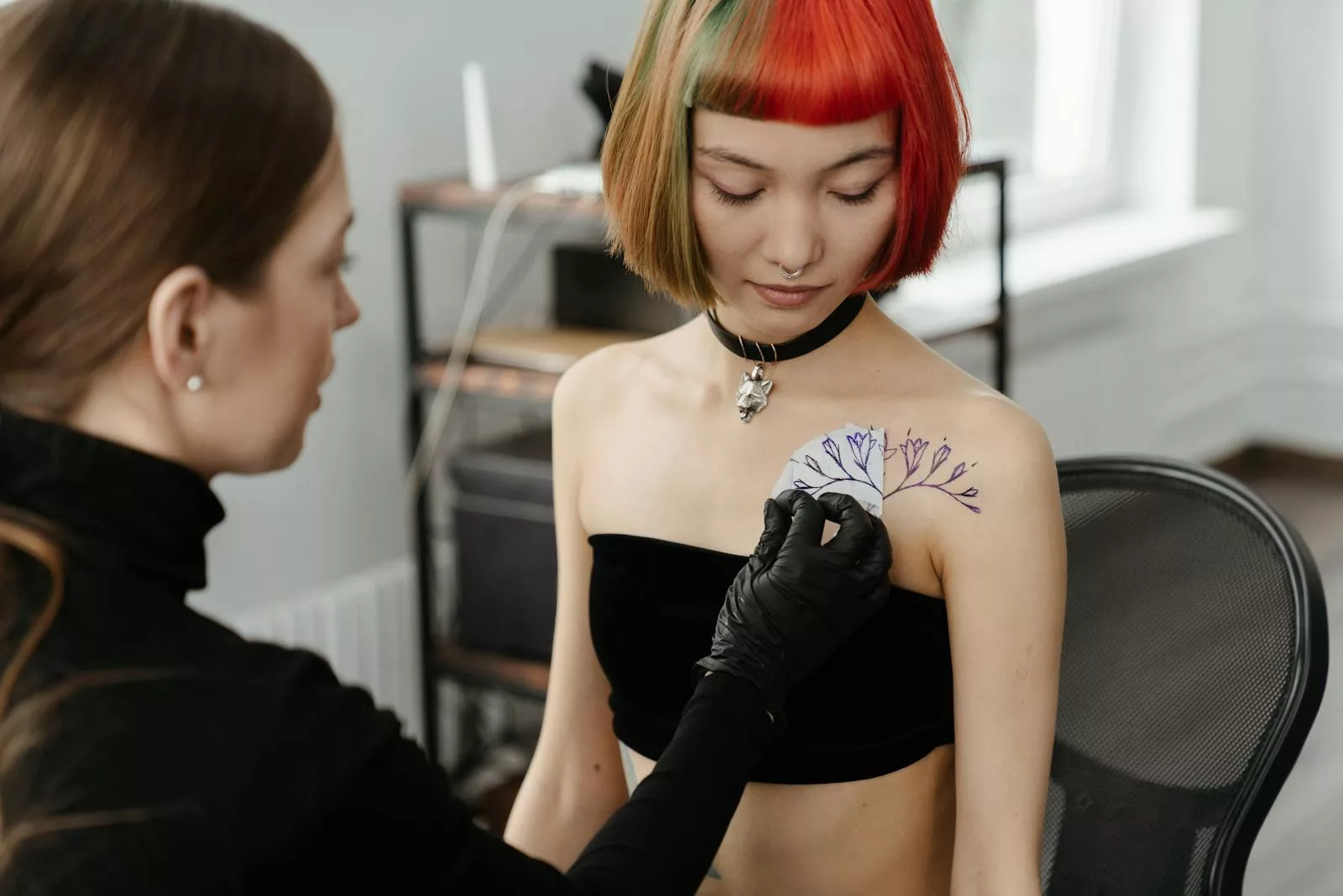 A tattoo artist applies a floral design on a woman's shoulder in a studio, showcasing artistry and style.