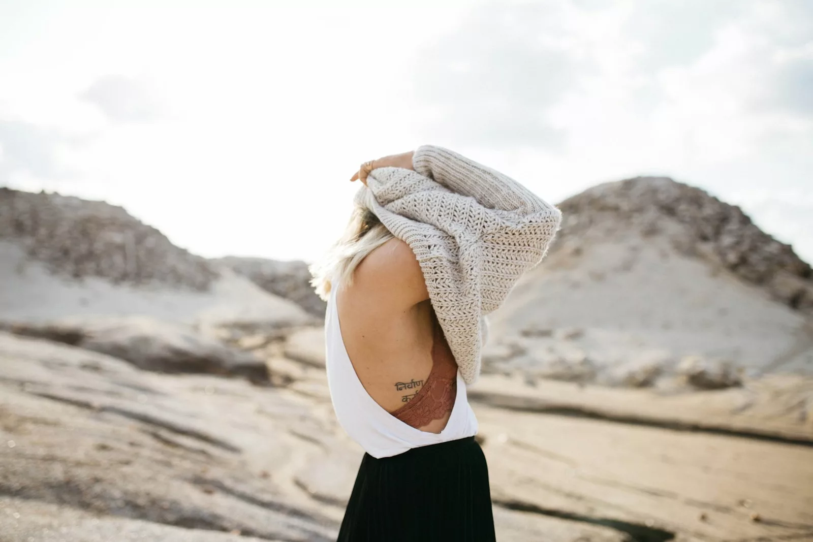 woman with rip tattoo about to wear beige knit sweater under white cloudy sky