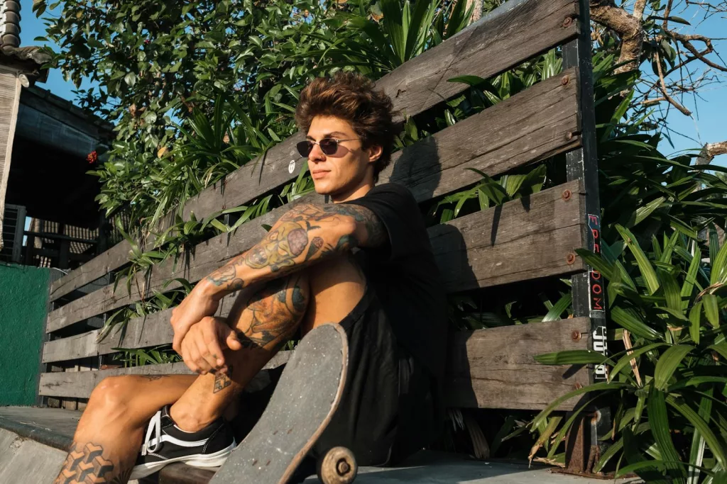 Skateboarder with tattoos relaxes on a sunny day in a park, wearing sunglasses and casual attire.