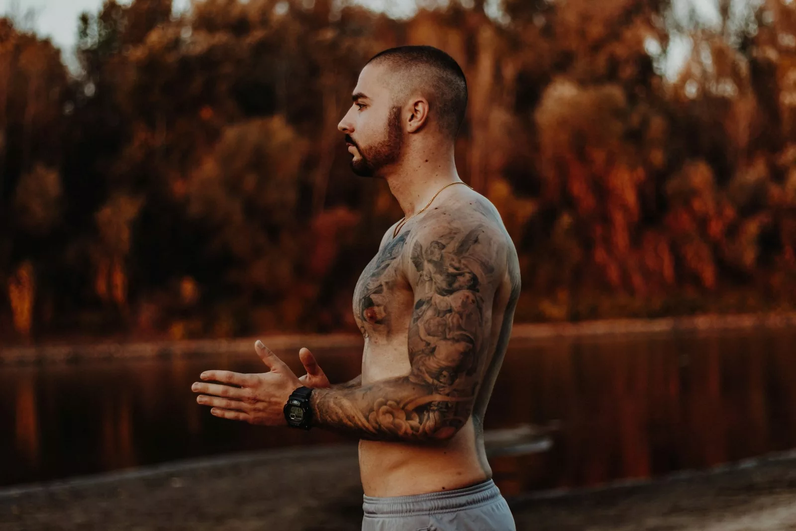 man wearing gray shorts standing in front of a lake. He has tattoos on his chest and arm.