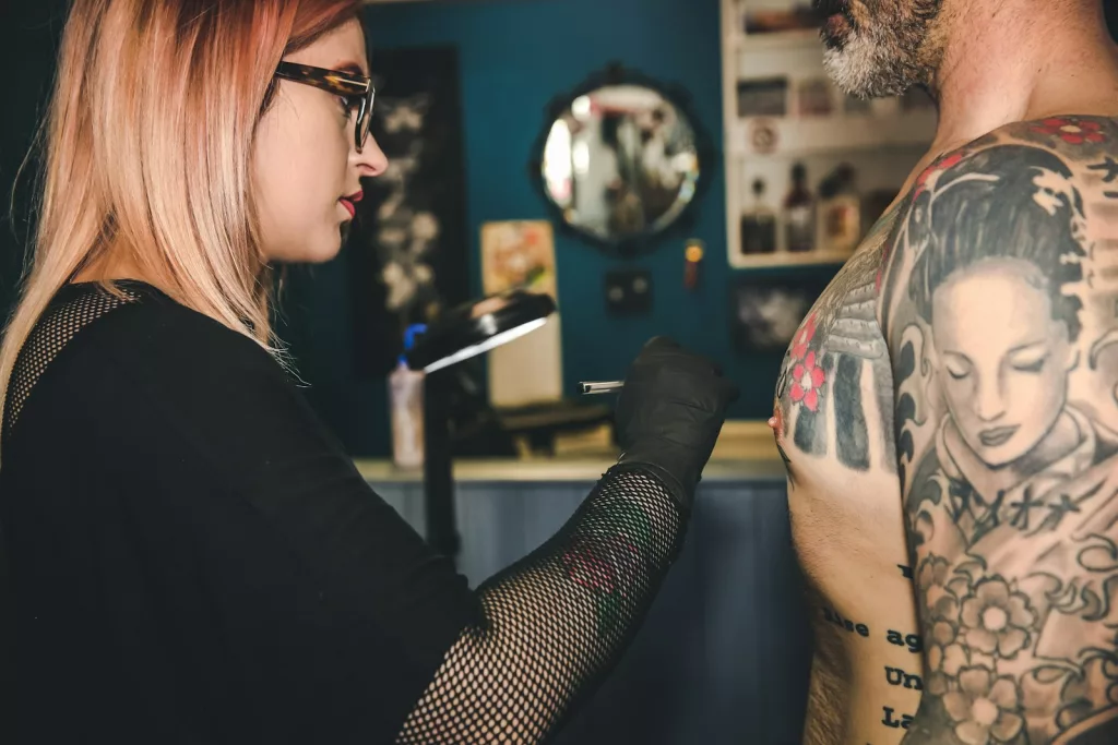 woman in black top is preparing to tattoo a man’s chest.