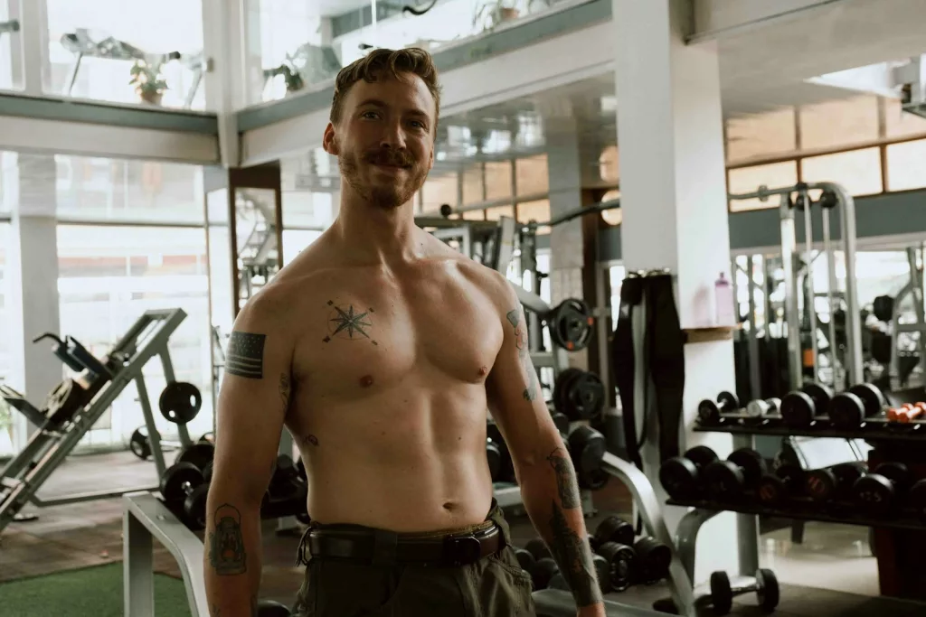 a shirtless man with tattoos on his chest and arm standing in a gym