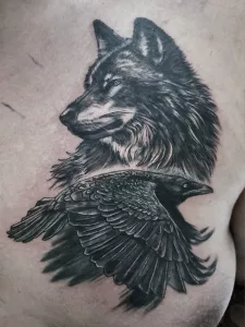 Nahaufnahme eines Tattoos auf der Brust. Das Tattoo zeigt einen fliegenden Raben und einen Wolf im Profil.