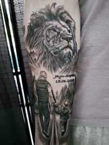Nahaufnahme eines Unterarm-Tattoos. Das Tattoo zeigt einen Löwenkopf und eine Person, die ein kleines Kind an der Hand führt.