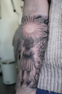 Nahaufnahme eines Unterarm-Tattoos in Black and Grey. Ein Mädchen mit Teddy in der Hand vor einem Sonnenuntergang.