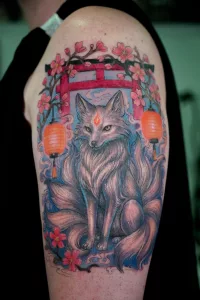 Nahaufnahme eines farbigen Oberarm-Tattoos von einem japanischen Kitsune vor Laternen und Torii.