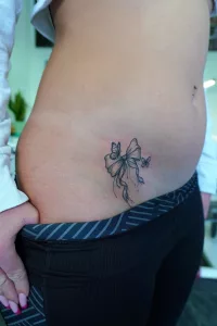 Nahaufnahme eines zierlichen Tattoos an der Hüfte: eine gebundene Schleife, um die zwei kleine Schmetterlinge flattern. Black-and-Grey-Stil.
