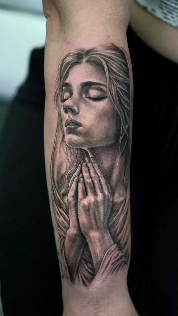 Nahaufnahme eines Unterarm-Tattoos. Das Tattoo zeigt das Porträt einer betenden Frau in black and grey.