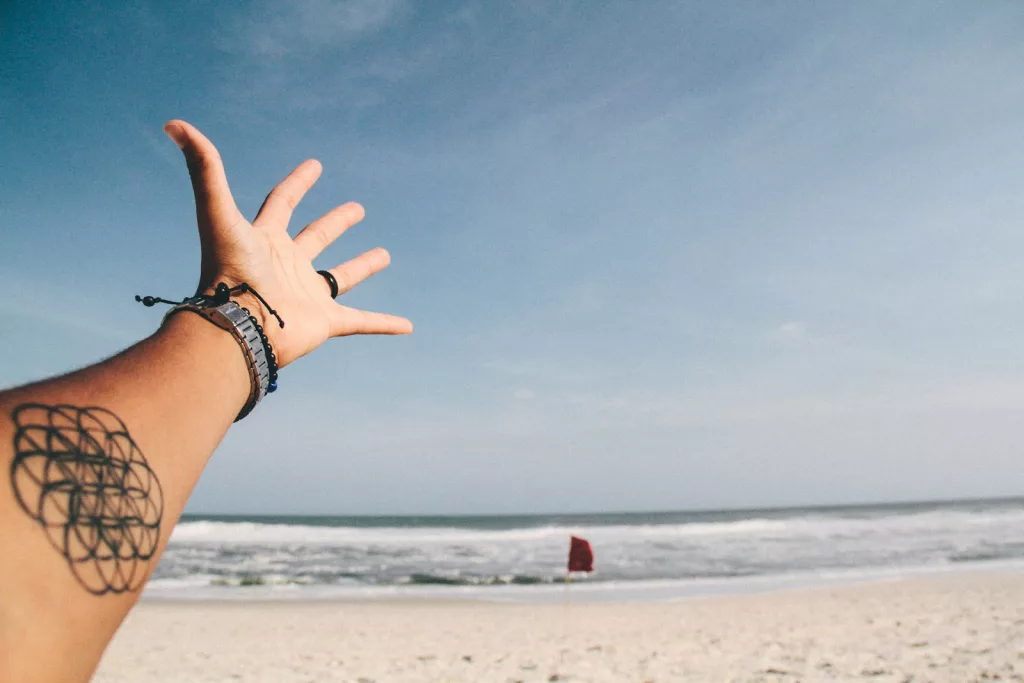 person raising it's hand near shoreline. On the arm is a geometric tattoo.