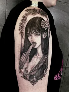 Nahaufnahme eines Oberarm-Tattoos in Black and Grey. Das Motiv zeigt einen asiatisches Mädchen mit langen Haaren, das die Zunge an ein Messer hält.