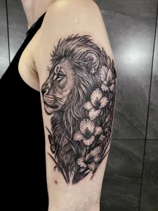 Nahaufnahme eines Oberarm-Tattoos in Black and Grey. Das Motiv zeigt einen Löwen im Profil, Blumen ranken sich seitlich an seiner Mähne entlang.