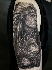 Nahaufnahme eines realistischen Oberarm-Tattoos in Black and Grey. Das Motiv zeigt einen Indianerhäuptling mit Kopfschmuck, der einen Wolf im Arm hält.