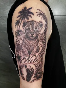 Nahaufnahme eines Oberarm-Tattoos in Black and Grey. Das Motiv zeigt einen Baby-Löwen, im Hintergrund sind Wellen und Palmen zu sehen, im Vordergrund ist ein Babyfußabdruck und ein Namensschriftzug.