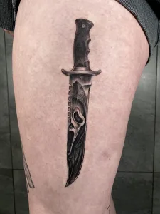 Nahaufnahme eines Oberschenkel-Tattoos in Black and Grey. Das Motiv zeigt einen Dolch, in dessen Klinge sich der Maskierte aus Scream spiegelt.