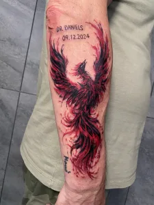 Nahaufnahme eines Unterarm-Tattoos in farbig. Das Motiv zeigt einen aufsteigenden Phönix in schwarz und tiefrot zusammen mit einem kleinen Namensschriftzug plus Datum.