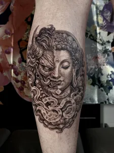 Nahaufnahme eines Unterschenkel-Tattoos in Black and Grey. Das Motiv zeigt einen Buddha und einen Dämon jeweils im halben Porträt, wobei die Gesichter ineinander übergehen.